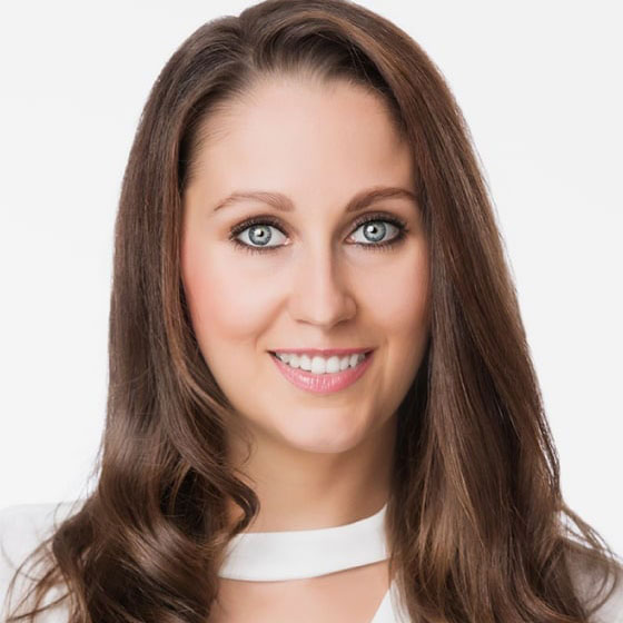 The image features a woman with long brown hair smiling at the camera  she has blue eyes and is wearing a white top with a neckline.