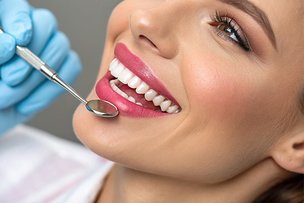 A woman with a broad smile receiving dental treatment, as she sits under a dental light and has her teeth cleaned by a professional using a dental tool.