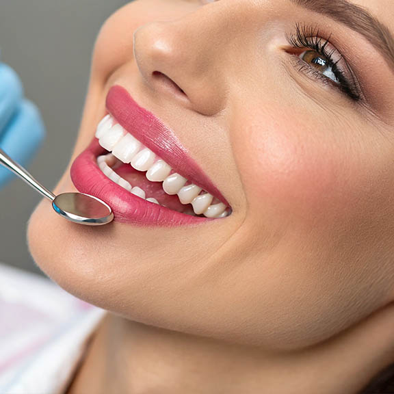 The image shows a smiling person with a bright smile, sitting in front of a dental professional who appears to be performing some form of oral care procedure, possibly related to teeth whitening, given the presence of a tray with a blue substance and a small brush-like tool.