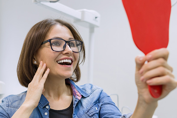 A woman with glasses, smiling, holding a red spatula next to her face while standing in front of a mirror.