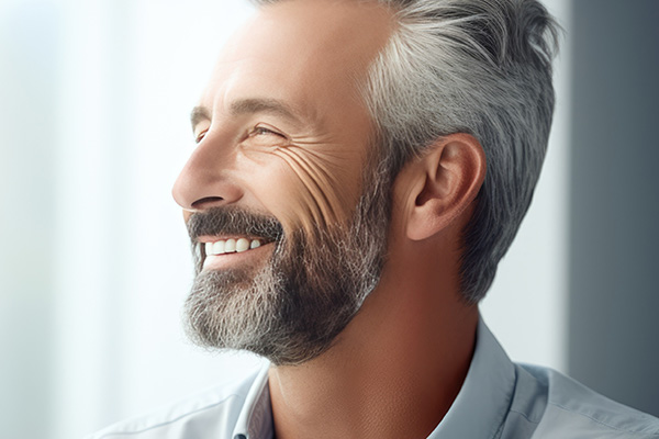 The image shows a man with gray hair and a beard, smiling at the camera, wearing a suit and tie, standing against a blurred background.