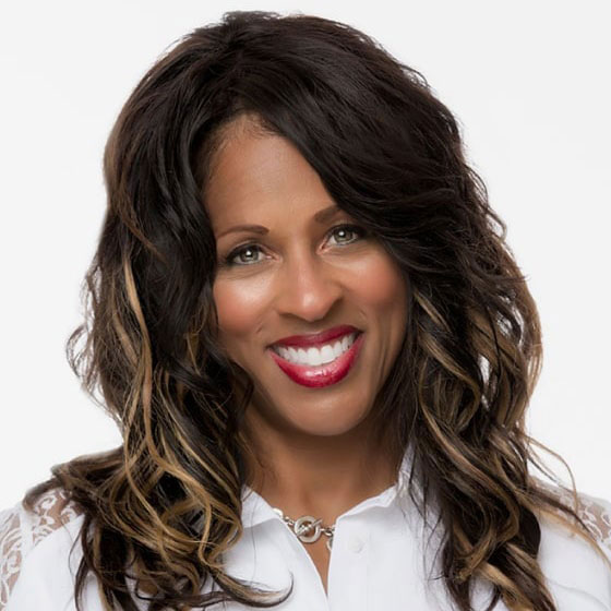 The image features a woman with long hair, wearing a white top, smiling at the camera, with her teeth visible. She has a red lipstick on and appears to be indoors with a plain background.