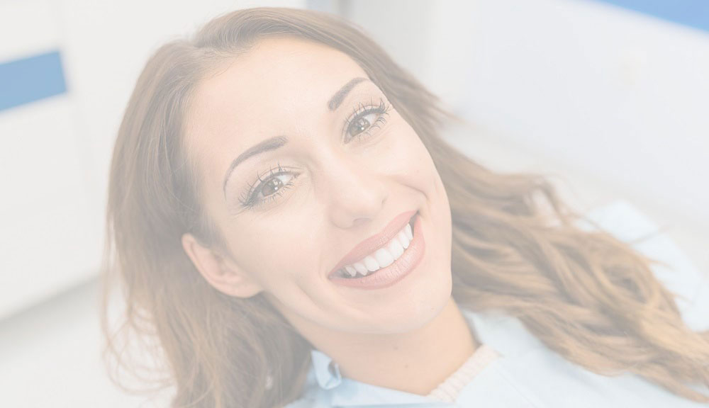 The image shows a smiling woman with long hair sitting in front of a blue background  she appears to be in a dental or medical setting, indicated by the presence of a chair and a sign with text that is too small to read clearly.