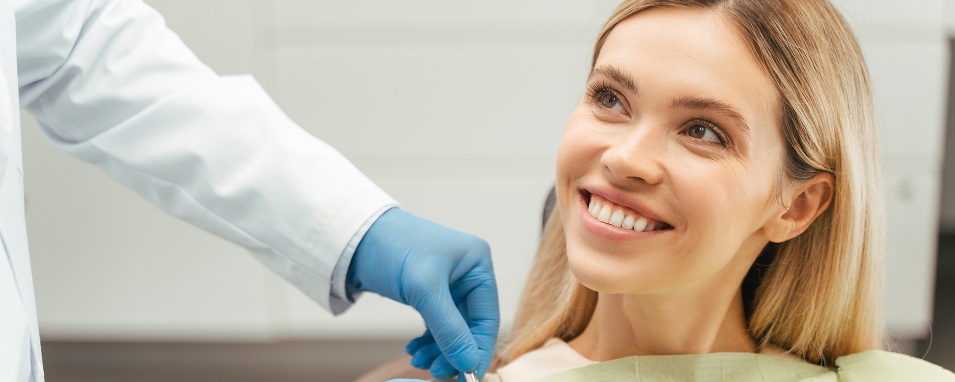 The image shows a woman seated in a dental chair being attended to by a dentist who is wearing a white coat, blue gloves, and a surgical mask.