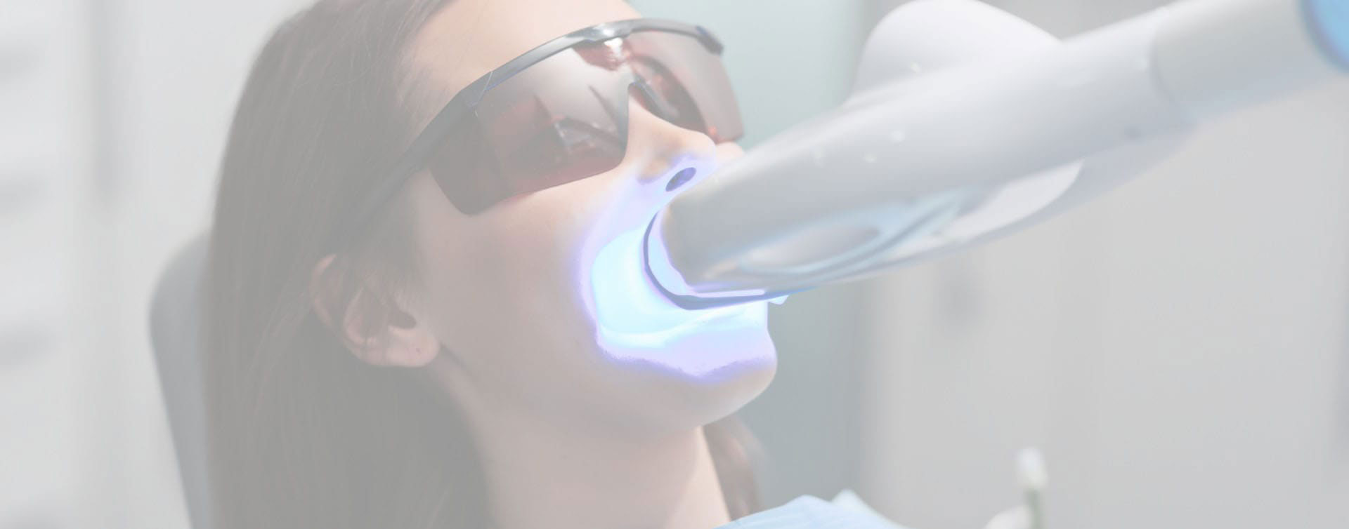 The image shows a person sitting in a dental chair with a blue light device on their face, likely for teeth whitening or cleaning purposes.