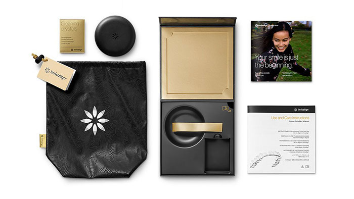 The image displays a collection of items neatly arranged on a white background, including a gold box with a black lid containing a product, a small bag with a tag that reads  THE BEGINNING,  a black envelope with a white label and a gold seal, a black and white booklet with a cover featuring a person s face, a black and white brochure with text, and a black and white pamphlet with a design on the front. The items are presented in an organized manner, suggesting they may be part of a product package or promotional material for a brand or event.
