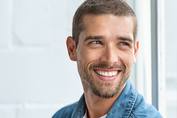 The image shows a man with short hair smiling at the camera. He appears to be indoors, possibly in an office environment, given the presence of blinds in the background.