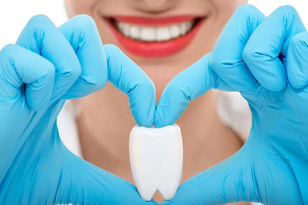 The image shows a person wearing blue gloves and holding up two fingers with a heart shape formed by their fingers. They are smiling and appear to be making a positive gesture, possibly related to dental care or hygiene given the presence of a toothbrush in their hand.