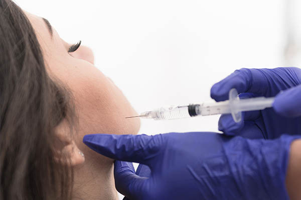 The image depicts a person receiving an injection with a syringe held by someone wearing blue gloves and a medical mask, set against a blurred background.