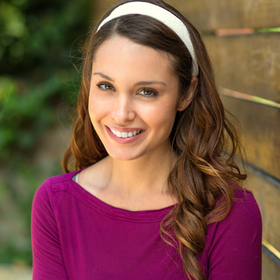The image features a young woman with long brown hair wearing a purple top and a white headband, smiling at the camera while standing against a wooden fence.