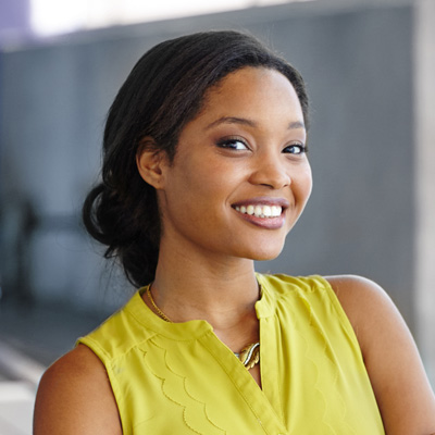 A smiling woman with dark hair wearing a yellow top and standing against a blurred background.